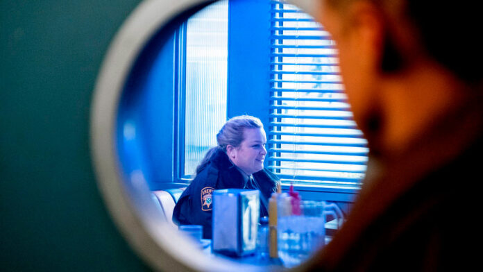 screenshot of deputy liv sitting drinking coffee at the diner, all seen through a circular window at the door.