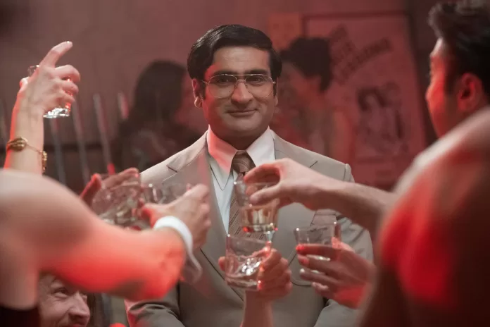 Photo of Kumail Nanjiani as Steve Banerjee awkwardly standing in a suit with a strong drink in hand, toasting with a series of white arms.